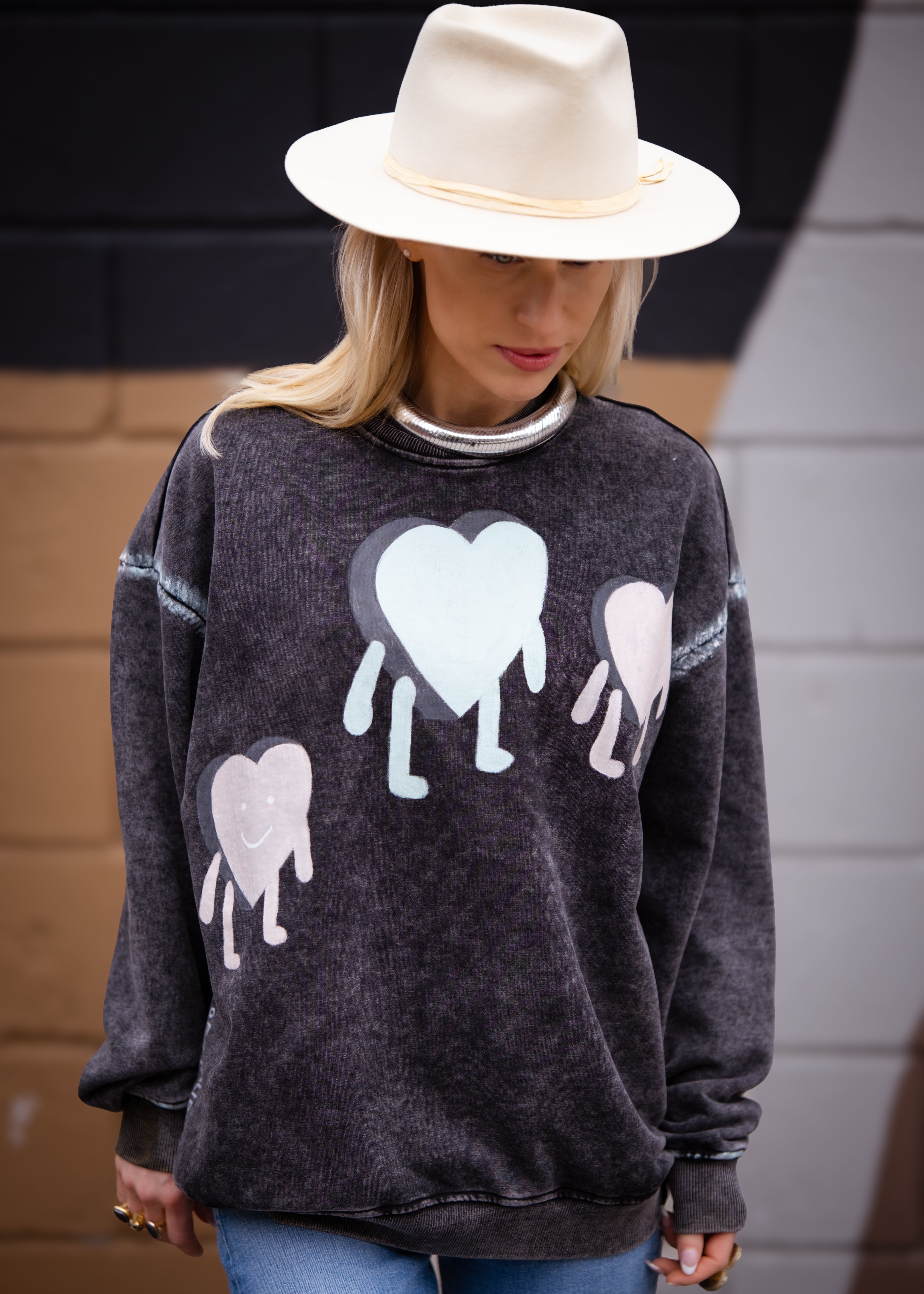 A woman wearing a washed black crewneck sweatshirt with hand-painted heart figures and text on the front, paired with a white hat and blue jeans.